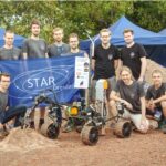 a group of people next to a rover on dirt ground, holding a STAR Dresden banner