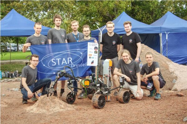 a group of people next to a rover on dirt ground, holding a STAR Dresden banner