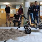 a rover driving on a sand pile indoors, multiple people watching