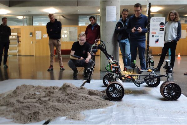 a rover driving on a sand pile indoors, multiple people watching